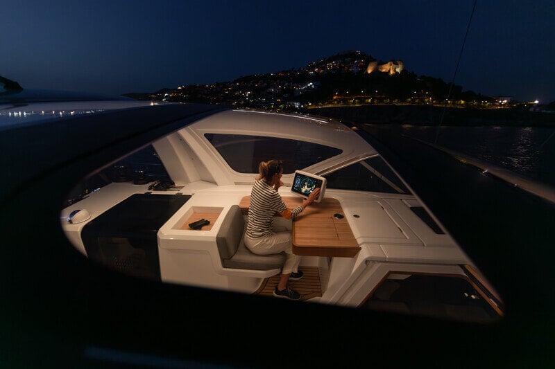 Nainen navigoi Jeanneau Yachts 55:n suojatussa ohjaamossa yöllä, käyttäen modernia navigointijärjestelmää valaistun rannikon loisteessa.