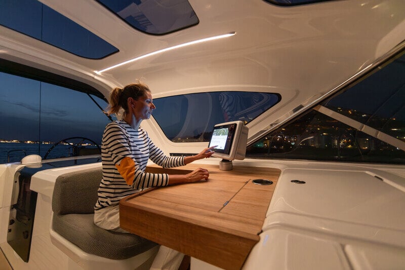 Nainen istuu Jeanneau Yachts 55:n suojatussa ohjaamossa yöllä ja käyttää navigointijärjestelmää valaistun rannikon edessä.